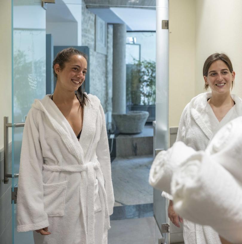 Due donne sorridenti in accappatoio bianco in un ambiente spa rilassante.